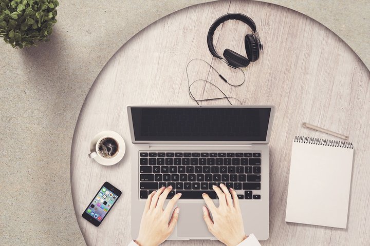 Rankos, dirbančios kompiuteriu, šalia telefonas, ausinės ir kavos puodelis